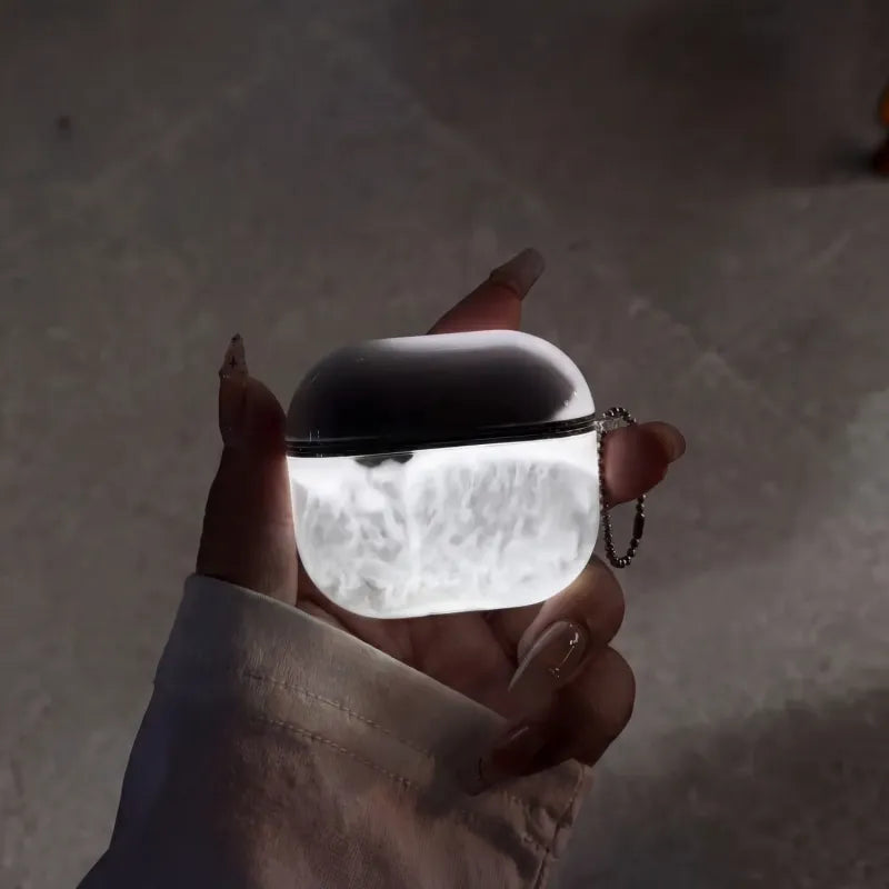 Midnight Smoke LumiCases AirPods cover glowing white beneath dark smoke marbling, held in a hand with its keychain